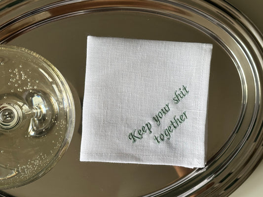 Lniana chusteczka z napisem „keep your shit together” na metalowej tacy, obok kieliszka z szampanem. 