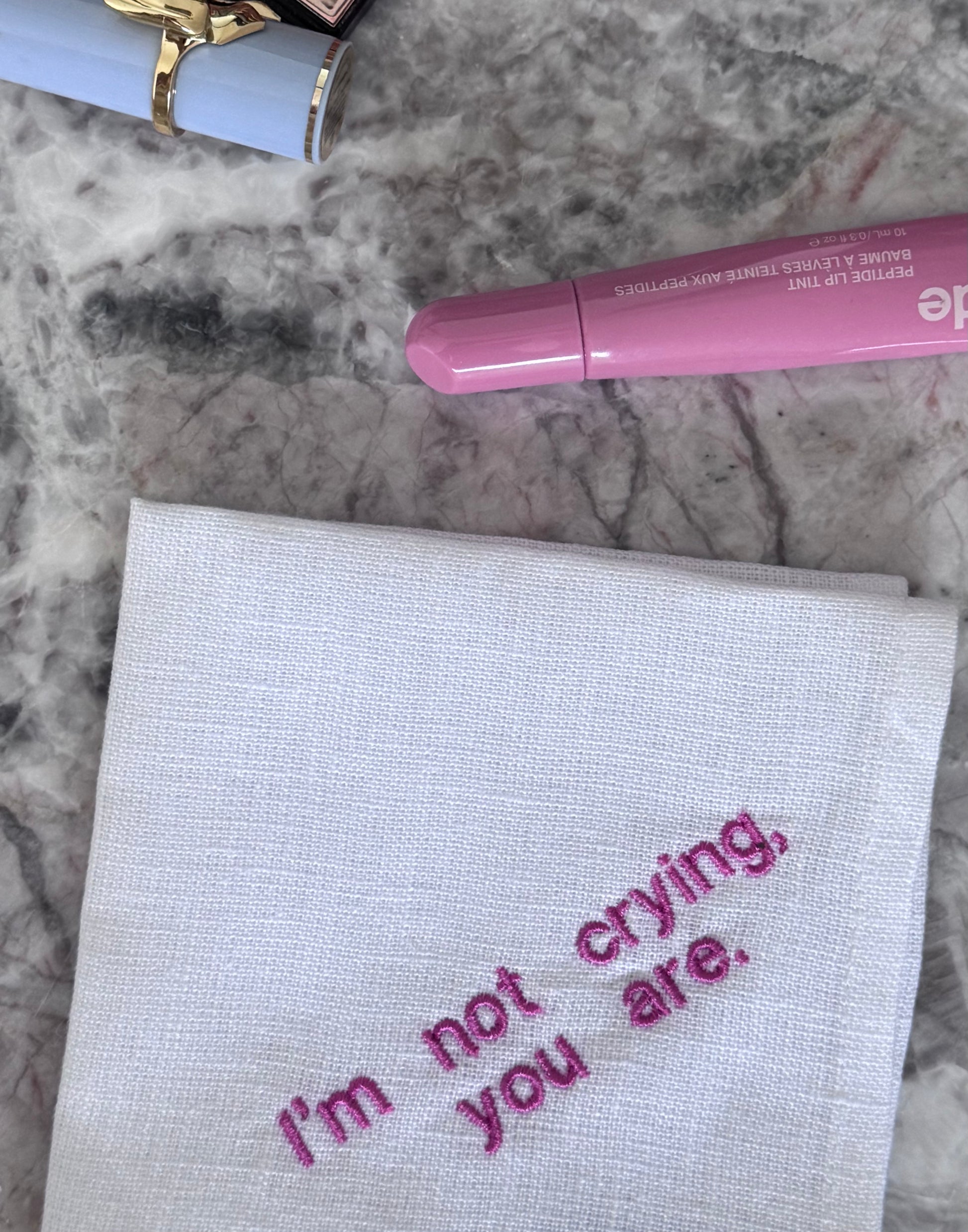 Chusteczka z napisem w kolorze różowym „I'm not crying, you are”, obok niej leży błyszczyk i szminka na marmurowym podłożu.